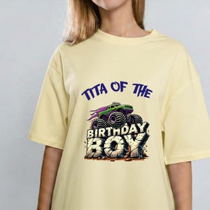 May include: A light yellow t-shirt with the text "TITA OF THE BIRTHDAY BOY" in blue and a graphic of a green monster truck with purple flames. The shirt is a casual style.