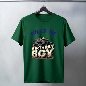 May include: A green t-shirt featuring a graphic of a monster truck with the text "TITA OF THE BIRTHDAY BOY" in purple and white. The monster truck is purple and green with large black tires. The text "BIRTHDAY BOY" is in a stone-like font.