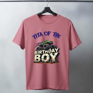 May include: A dusty rose t-shirt featuring a graphic of a green monster truck with purple accents, atop the words "BIRTHDAY BOY" in a stone-like font. Above the truck, the text reads "TITA OF THE" in blue.