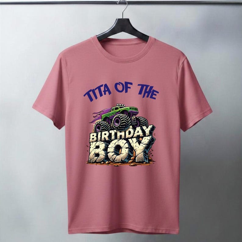 May include: A dusty rose t-shirt featuring a graphic of a green monster truck with purple accents, atop the words "BIRTHDAY BOY" in a stone-like font. Above the truck, the text reads "TITA OF THE" in blue.
