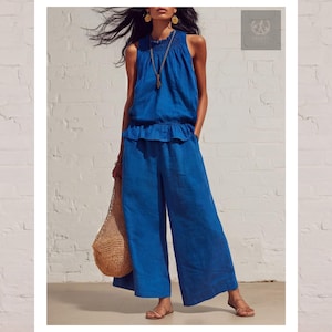 May include: A sleeveless, cobalt blue jumpsuit with wide legs. The top has a ruffled waist and a high neckline. A woven bag is held in one hand. The background is a white brick wall. The outfit is paired with gold sandals and jewelry.