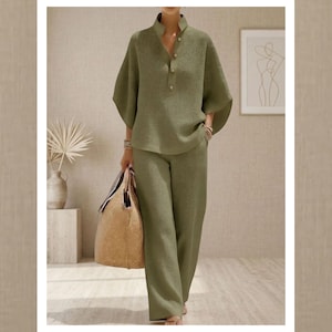 May include: A two-piece olive green outfit with a textured waffle weave. The top has a mandarin collar and button closure. The pants are wide-legged. A beige tote bag is carried. The background includes a framed art print and a vase with dried plants.