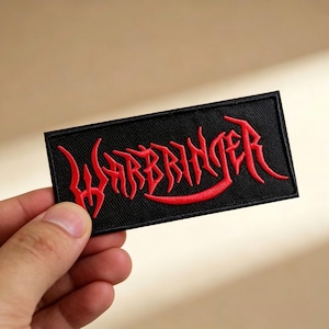 May include: Black rectangular patch with the word "WARBRINGER" embroidered in red, stylized lettering. The patch is held in a hand against a neutral background. The patch is likely for clothing or accessories.