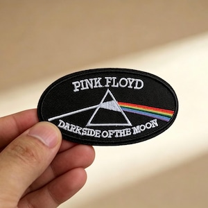 May include: Black oval patch with white embroidered text "PINK FLOYD" above a white triangle and rainbow. Below the triangle is the text "DARK SIDE OF THE MOON". The patch is held by a hand.