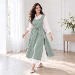 May include: A sage green sleeveless apron dress featuring a wide scoop neckline, a fitted bodice with a tie-waist belt, and a long, flowing skirt with side slits. The apron is layered over a long-sleeved white shirt and a matching white floor-length skirt.