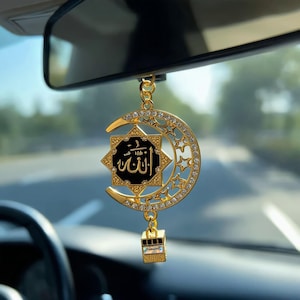 May include: A gold-toned car rearview mirror hanging ornament featuring a crescent moon and star motif encrusted with clear rhinestones. A central black square plaque displays gold Arabic calligraphy, and a small cube charm hangs below.