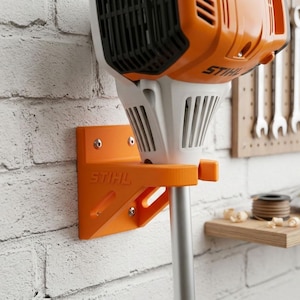 May include: An orange wall-mounted bracket with the STIHL logo embossed on the side, securely holding a white and orange STIHL power tool. The bracket is attached to a white brick wall with visible metal screws, providing a stable storage solution.
