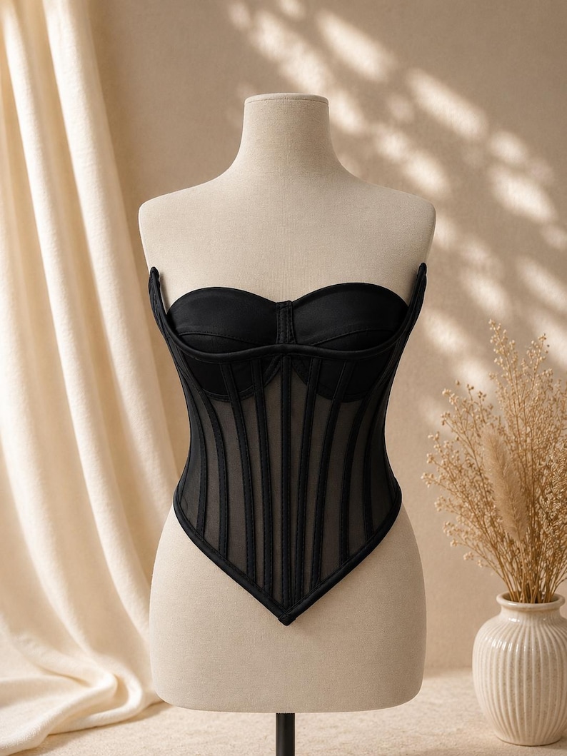 May include: A black strapless corset top featuring a sweetheart neckline, structured cups, and vertical boning channels. The bodice is constructed with sheer mesh panels between the opaque fabric strips, ending in a pointed hemline at the waist.