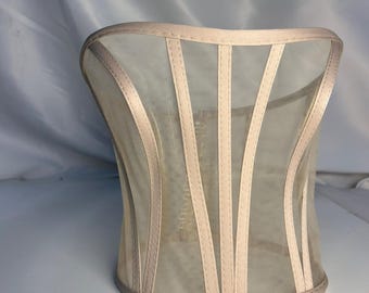 May include: A beige corset top constructed from sheer mesh fabric, featuring vertical satin-finish boning channels that run from the top edge to the bottom hem. The garment has a straight neckline and a structured, form-fitting silhouette.