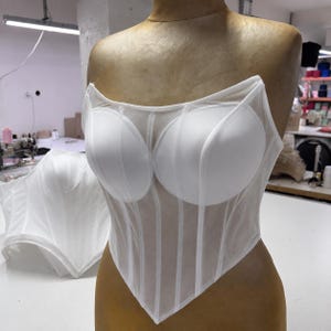 May include: A white strapless corset top featuring structured, opaque molded cups and a sheer, semi-transparent mesh bodice with vertical boning channels. The garment has a pointed, V-shaped hemline and is displayed on a mannequin torso.