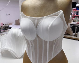 May include: A white strapless corset top featuring structured, opaque molded cups and a sheer, semi-transparent mesh bodice with vertical boning channels. The garment has a pointed, V-shaped hemline and is displayed on a mannequin torso.