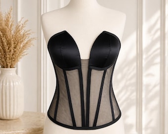 May include: A black strapless corset with a plunging neckline. The bodice features a sheer mesh panel with vertical black boning. The top is made of a shiny black fabric. The corset is displayed on a mannequin.