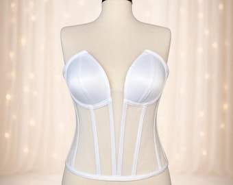 May include: A white strapless corset with a deep V-neckline. The bodice is made of satin and sheer panels, with vertical boning for structure. The corset is displayed on a mannequin against a soft, blurred background.