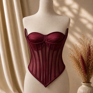 May include: A deep burgundy corset top featuring a sweetheart neckline, structured cups, and vertical boning channels. The bodice is constructed with a combination of opaque satin panels and sheer mesh inserts, ending in a pointed hemline.
