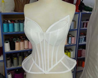 May include: A white strapless corset top featuring a sheer mesh bodice with vertical boning channels and a structured peplum hem. The garment has a sweetheart neckline with opaque molded cups and is displayed on a beige dress form mannequin.