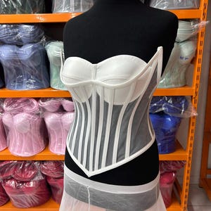 May include: A white strapless corset top featuring structured cups and vertical boning channels. The bodice is constructed with a combination of opaque white panels and sheer mesh inserts, creating a defined silhouette with a pointed hemline at the waist.