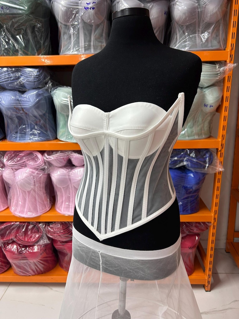 May include: A white strapless corset top featuring structured cups and vertical boning channels. The bodice is constructed with a combination of opaque white panels and sheer mesh inserts, creating a defined silhouette with a pointed hemline at the waist.
