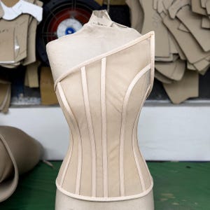 May include: A beige corset top featuring a single shoulder strap design, constructed with vertical boning channels and sheer mesh panels. The garment is displayed on a mannequin, showcasing a structured silhouette with light-colored piping.