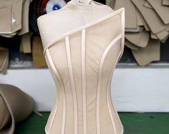 May include: A beige corset top featuring a single shoulder strap design, constructed with vertical boning channels and sheer mesh panels. The garment is displayed on a mannequin, showcasing a structured silhouette with light-colored piping.
