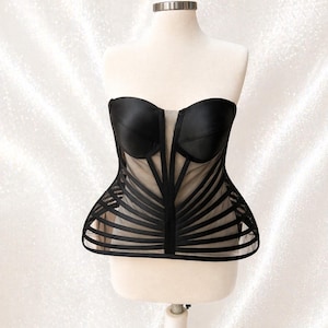 May include: A black strapless corset top featuring structured satin cups and a sheer mesh bodice. The garment is detailed with vertical and horizontal black satin ribbon overlays that create a cage-like effect over the nude-colored mesh base.