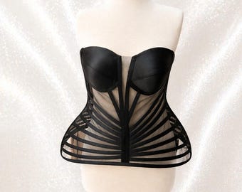 May include: A black strapless corset top featuring structured satin cups and a sheer mesh bodice. The garment is detailed with vertical and horizontal black satin ribbon overlays that create a cage-like effect over the nude-colored mesh base.