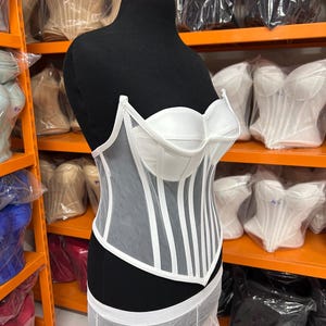 May include: A white corset top featuring structured cups and vertical boning channels. The garment is constructed with a combination of opaque white fabric panels and sheer white mesh panels, creating a layered look with a pointed hemline.