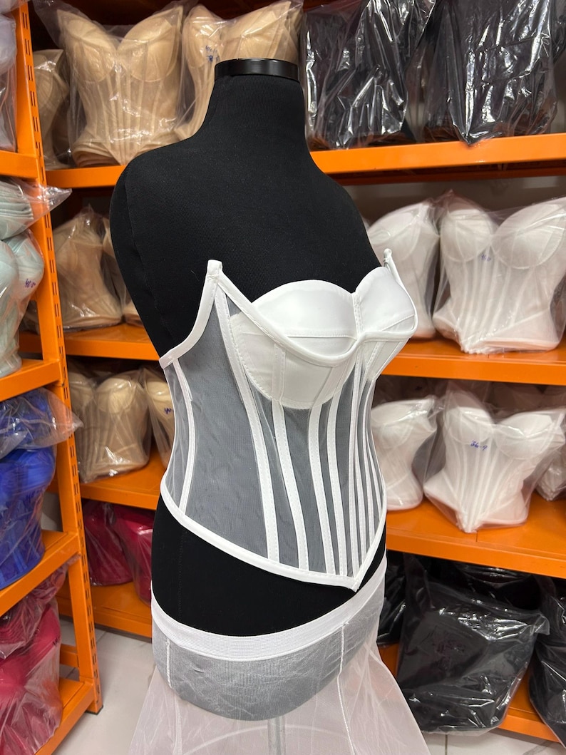May include: A white corset top featuring structured cups and vertical boning channels. The garment is constructed with a combination of opaque white fabric panels and sheer white mesh panels, creating a layered look with a pointed hemline.