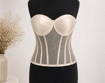 May include: A beige strapless corset displayed on a black mannequin. The corset features a structured bust with vertical boning and a sheer mesh panel. The design includes beige trim and a smooth, satin-like fabric.
