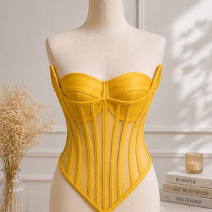 May include: A bright yellow strapless corset top featuring a sweetheart neckline, vertical boning channels, and sheer mesh panels between the structured fabric sections. The garment has a pointed hemline that dips into a V-shape at the center front.