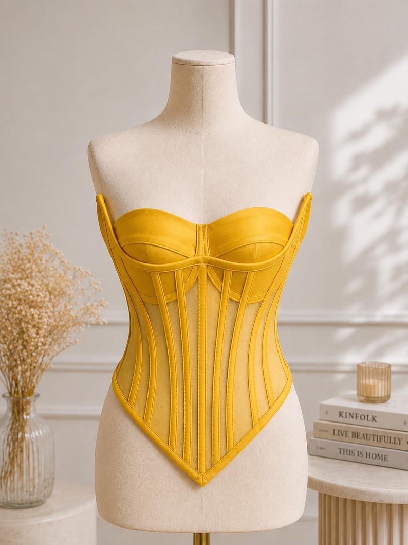 May include: A bright yellow strapless corset top featuring a sweetheart neckline, vertical boning channels, and sheer mesh panels between the structured fabric sections. The garment has a pointed hemline that dips into a V-shape at the center front.
