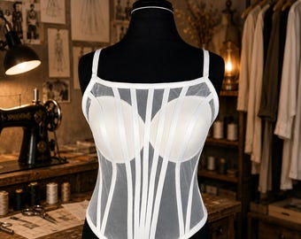 May include: A white corset top with a sheer overlay and vertical stripe detailing. The bustier features padded cups and thin shoulder straps. The garment is displayed on a black mannequin in a studio setting.