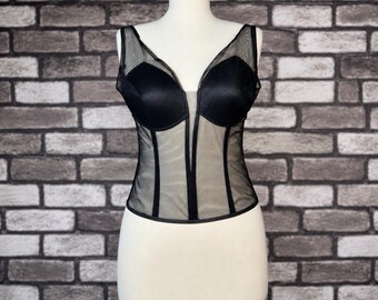 May include: A black corset top featuring opaque molded cups and a sheer mesh bodice. The garment includes a deep V-neckline, vertical boning details, and a structured silhouette, displayed on a mannequin against a textured brick wall background.