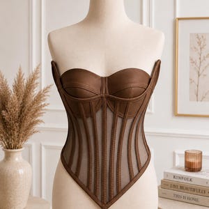 May include: A strapless brown corset top featuring structured cups and vertical boning channels. The bodice is constructed with opaque brown fabric panels alternating with sheer mesh inserts, creating a defined silhouette with a pointed hemline.