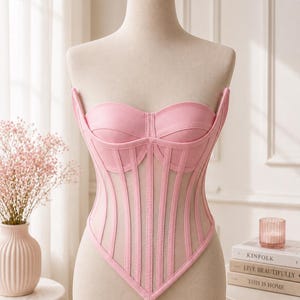 May include: A light pink strapless corset top featuring a sweetheart neckline, structured boning, and sheer mesh panels between the vertical fabric strips. The garment has a pointed hemline and a fitted silhouette displayed on a mannequin.