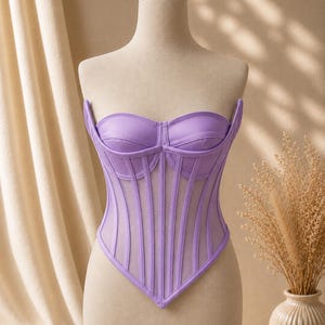 May include: A lavender corset top featuring a sweetheart neckline, structured cups, and vertical boning details. The garment is constructed with a combination of solid lavender fabric panels and sheer mesh inserts, creating a pointed hemline.