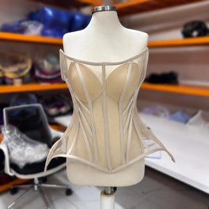 May include: A strapless beige corset top featuring a sheer mesh bodice with structured boning, molded cups, and a pointed hemline. The garment is displayed on a mannequin, showcasing vertical fabric panels and a fitted, architectural silhouette.