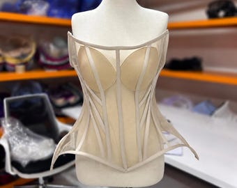 May include: A strapless beige corset top featuring a sheer mesh bodice with structured boning, molded cups, and a pointed hemline. The garment is displayed on a mannequin, showcasing vertical fabric panels and a fitted, architectural silhouette.