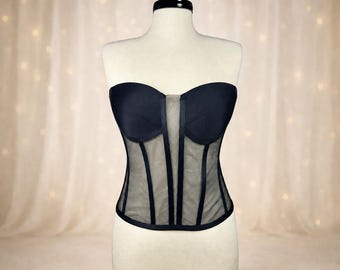 May include: A black strapless corset displayed on a mannequin. The corset features a sheer mesh panel in the center and vertical boning details. The bust is made of a solid black fabric.