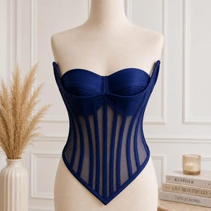May include: A strapless navy blue corset top featuring a sweetheart neckline, structured cups, and vertical boning channels layered over sheer mesh panels. The bodice has a pointed hemline that dips to a center point at the waist.
