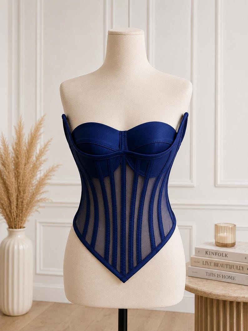 May include: A strapless navy blue corset top featuring a sweetheart neckline, structured cups, and vertical boning channels layered over sheer mesh panels. The bodice has a pointed hemline that dips to a center point at the waist.