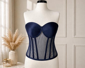 May include: A navy blue strapless corset displayed on a mannequin. The corset features a sweetheart neckline, sheer mesh paneling, and vertical boning details. The garment is a classic lingerie piece.