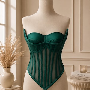 May include: A dark green strapless corset top featuring a sweetheart neckline, structured boning, and vertical sheer mesh panels. The garment has a pointed hemline and a solid opaque fabric finish on the bust and along the vertical seams.
