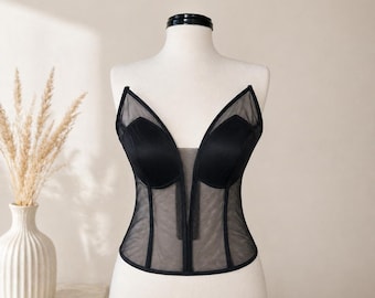 May include: A black corset with a deep V-neckline and sheer mesh panels. The bustier is displayed on a mannequin, showcasing its structured design and delicate straps. The garment is a lingerie piece.