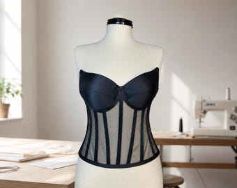 May include: A black strapless corset top featuring molded cups and a sheer mesh bodice with vertical boning channels. The garment has a sweetheart neckline and a structured, fitted silhouette that extends to the waist, showcasing a clean design.