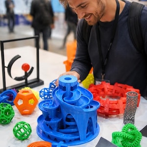 May include: A collection of 3D printed objects including a large blue spiral marble run, a bright orange dodecahedron, several green and blue geometric lattice spheres, a red interlocking ring structure, and a grey cylindrical mesh tube.