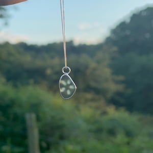 Könnte beinhalten: Eine Silberkette mit einem kleinen, unregelmäßig geformten, durchscheinenden Anhänger, in den ein weißes, sechsblättriges Blumenmuster eingebettet ist. Der Anhänger hängt an einer feinen Silberkette und glänzt an seinem polierten Rand.