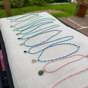 Könnte beinhalten: Eine Sammlung von Perlenketten, die in einer Reihe angeordnet sind und jeweils einen kleinen, glatten, kieselartigen Anhänger aufweisen. Die Perlen variieren in der Farbe, einschließlich Grün-, Blau- und Rosatönen, mit einem silberfarbenen Verschluss.