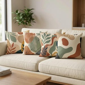 May include: Four decorative throw pillows with abstract botanical designs in shades of green, orange, and brown on a cream background. The pillows are arranged on a beige sofa, creating a cozy and stylish living room setting.