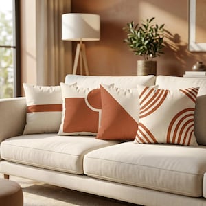 Pode incluir: Quatro almofadas quadradas dispostas num sofá bege claro. Cada almofada apresenta uma base de cor creme com padrões geométricos distintos em terracota, incluindo uma faixa horizontal, um quadrado com um círculo, um triângulo grande e arcos concêntricos.