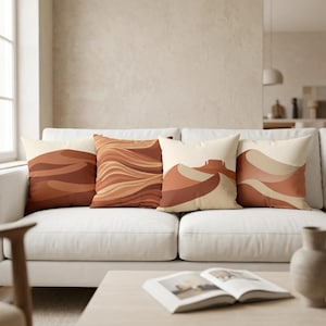 May include: Four square throw pillows arranged on a white sofa. Each pillow features a distinct desert-inspired graphic print in shades of terracotta, burnt orange, cream, and beige, showcasing abstract wavy lines and stylized landscape formations.